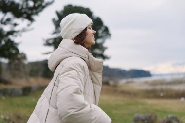 Şık bir kış yolcusunun portresi kabarık bir ceket ve şapkayla açık havada sakin bir kırsal alan, uzak bir ufuk ve sessiz bir gökyüzünün altında hafif bir esinti.