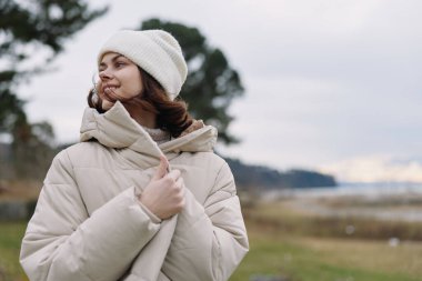 Açık havada kabarık bir ceket ve örgü şapka giymiş bir kadının portresi, rüzgarla savrulan saçlar, dingin bir ifade, serin havada mevsimsel stil ve uzak bir gökyüzü.