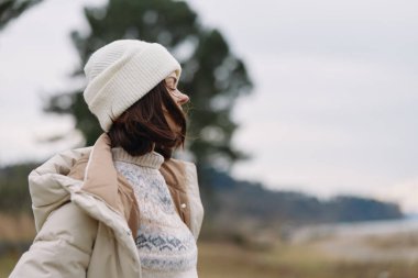 Sıcak ceketli, atkı ve şapkalı, soğuk bir ortamda, rüzgârlı bir havada, sakin kırsal ve kış tarzında bir kadınla açık hava sahnesi..