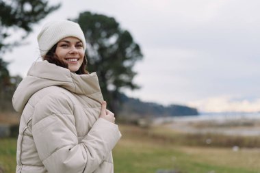 Kışlık palto giyen ve bereli gülümseyen bir kadın rüzgarlı bir tarlada duruyor. Rahatlamış özgüveni, açık hava yaşam tarzını ve gri gökyüzüne karşı mevsimsel sıcaklığı tasvir ediyor.