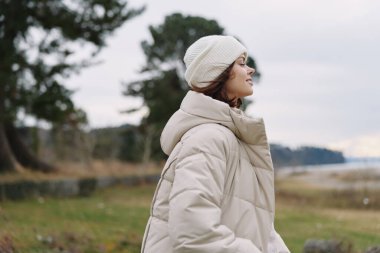 Açık hava sahnesinde, gölün kenarında bej ceketli ve bereli bir kadın, sıcaklığı, rahatlığı ve doğadan esinlenerek moda yaratıyor.