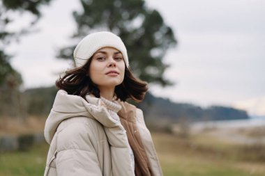 Havalı bir ceket ve örgü bir şapka giyen şık bir kadının açık hava portresi, rüzgarlı bir manzarada, su ve ağaçlarla, sakinliği, esnekliği ve mevsimselliği aktarıyor..