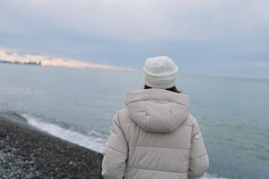 Rüzgarlı bir sahil şeridinde, kapüşonlu kışlık ceketli yalnız bir şahıs, uzak ufuk ve gri bulutlar ile sessiz denize bakıyor, sakin kıyılar ve yansıtıcı ruh hali..