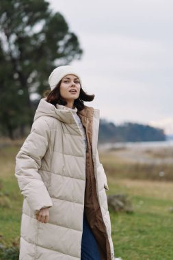 Açık hava moda sahnesi. Krem rengi kabarık ceketli ve örgü şapkalı bir kadın. Çimenli bir manzaranın yanında duruyor..