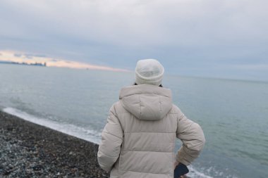 Kayalık bir plajda duran, okyanusu ve uzak kıyı şeridini izleyen, sakin bir yansımayı, soğuk hava havasını ve sakin bir kıyı manzarasını yansıtan bir şahsın arka görüntüsü..