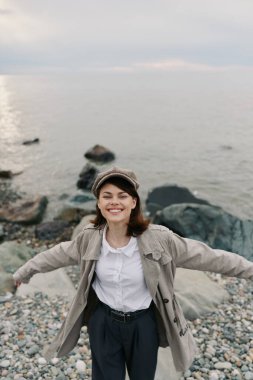 Açık renk ceketli ve şapkalı gülümseyen kadın kollarını çakıl taşı plajında taşlı huzurlu okyanus suyuyla açıyor. Mutluluk kavramı, rahatlama ve temiz havanın ve doğal manzaranın tadını çıkarma..