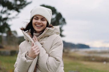 Serin bir manzarada ceketi ve atkısı olan gülümseyen bir kadının açık hava portresi. Samimi, rahat bir hava, rüzgarlı saçlar, yumuşak renkler ve sakin bir manzara sakin bir yaşam tarzı ve açık hava eğlencesi vurguluyor..