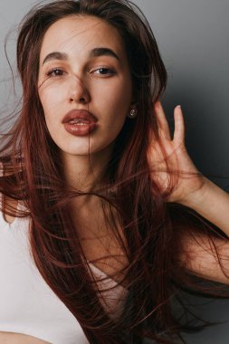 Dalgalı saçlı ve doğal görünümlü bir kadın portresi, el dokunmalı saçlar, beyaz bluz giyiyor, yüzünde hafif bir aydınlatma var, gri arka planda sakin bir ruh hali içinde izole edilmiş..