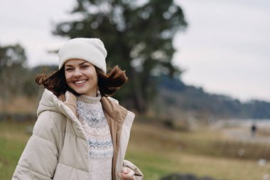 Sıcak bir palto ve örgü şapka içinde gülümseyen bir kadının açık hava portresi, çimen ve uzak ağaçlar ile sakin bir manzara, rahatlık, tarz ve mevsimsel ruh hali vurgulanıyor