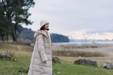 Sahilde bir kadın tarafından giyilen şık bir kışlık ceket, arka planda kum tepeleri ve su, sakin ruh hali, günlük moda ve doğa uyumu.