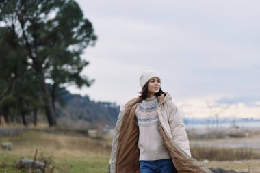 Açık hava sahnesinde rahat bir palto giymiş bir kadın, örgü bir eşarp ve göl kenarında bir bere. Rüzgarlı hava, dingin doğa ve huzurlu bir manzarada şık kış kıyafetleri..