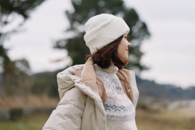 Açık hava sahnesinde, rahat örgü ve yumuşak ceketli bir kadın, beyaz bereli, sakin bir kış havası yakalıyor ve kırsal doğal bir ortamda şık bir tarz yakalıyor.