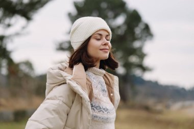 Kışlık ceketli ve örgü kazaklı bir kadının portresi, açık bir alanda narin bir gülümsemeyle duruyor, serin, pastoral bir çevrede huzurlu bir ruh hali ve doğal bir ışık yakalıyor.