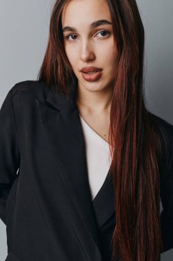 Uzun düz saçlı, siyah ceketli, beyaz gömlekli, sade arka planda duran, az makyajlı ve rahat duruşu olan kendine güvenen genç bir kadının portresi..