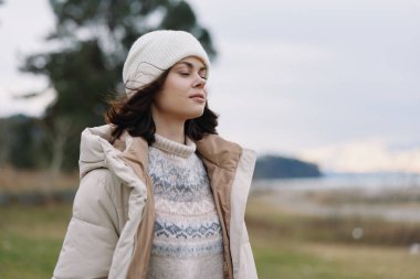 Kabarık bir ceket ve yün kazak giyen sakin bir kadının dış portresi, bereli ve rüzgarlı saçlar, açık alanlarda sakin bir ruh hali ve sakin bir kıyı şeridi, durgunluğu ve serin atmosferi yakalıyor..