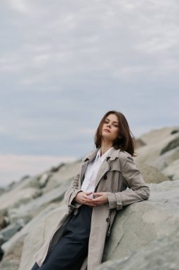 Uzun paltolu şık genç bir kadın bulutlu gökyüzünün altındaki kayalara yaslanıyor. Doğal ışık, sakin ve kayalık bir sahil ortamında gündelik tarz ve sakin ifadeyi vurgular.