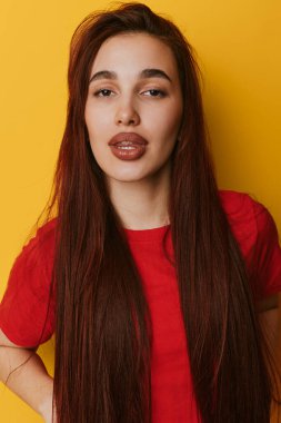 Uzun kahverengi saçlı ve kırmızı gömlekli genç bir kadın portresi. Sarı duvarın önünde duruyor. Doğal makyajlı ve rahat bir duruşu var..