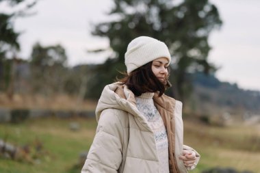 Açık kırsal kesimde kalın bir palto ve bere giyen bir kadının portresi, dingin bir ruh hali, serin hava modası, doğal sahne arkası, açık hava fotoğrafı..