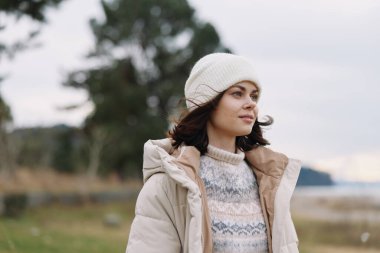 Açık hava portresi, yastıklı bir ceket ve yün şapka giyen bir kadın, yumuşak doğal ışık, açık arazi manzarası, dingin bir ruh hali ve yaşam tarzı ve moda stoku için rahat bir kış tarzı..