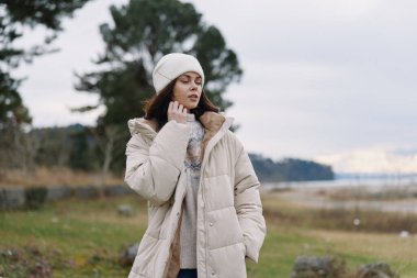 Sıcak bir palto ve örgü şapkalı şık bir kadının portresi, sakin bir kırsal alanda yumuşak bir ışık, sakin bir esneklik ve kış havasını yansıtıyor..