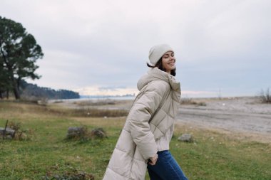 Sahilde uzun bir kabarık ceketli bir kadın, temiz havanın, deniz esintisinin ve yumuşak ışık altında sakin bir kışın tadını çıkarıyor..