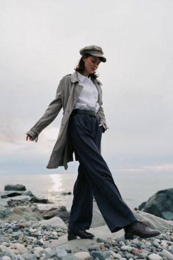 Uzun paltolu ve şapkalı şık genç bir kadın bulutlu arka planlı sakin deniz kenarında kayalık sahilde yürüyor. Tarz kavramı, sonbahar modası, açık hava yaşam tarzı, ve tabiatta kendine güvenen ruh hali..