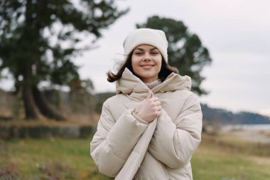 Rahat bir parka giymiş bir kadının açık hava portresi, örgü bir şapka ve atkı, dingin bir kış gününde ağaçlarla suyun kenarında duran, dingin bir ruh hali ve yumuşak bir ışık..