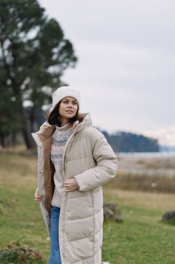 Kışlık ceketli, kabarık ceketli, kazaklı ve bereli bir kadınla açık hava sahnesi. Yumuşak bir gökyüzünün altında seyahat ve yaşam tarzı için moda ve doğa havası yaratıyor..