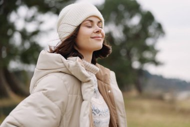 Açık hava portresi, rahat bej bir parka ve beyaz bir bere takan huzurlu bir ifade ve doğal bir manzarayla meltemin tadını çıkaran bir kadın..