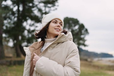 Sıcak bir ceket ve örgü bir şapka giyen, kırsal alanda temiz havanın tadını çıkaran, hafif ışıklı ve uzak tepeli bir kadının sıradan dış portresi..