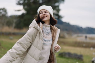 Kabarık paltolu ve bereli gülümseyen bir kadının açık hava portresi, yumuşak ışık ve doğal manzarayla geniş bir alanda duruyor..