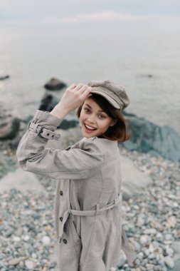 Bej ceketli ve şapkalı mutlu kadın çakıl taşı plajında su kenarında duruyor. Neşeli ve rahat görünüyor, doğal kıyı manzaralı açık hava sonbahar sahnesinden hoşlanıyor..