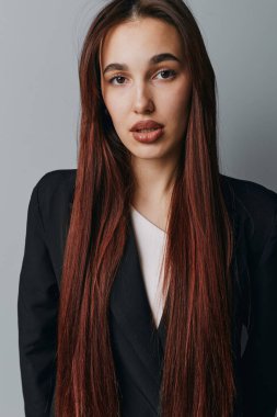 Siyah ceketli, uzun kahverengi saçlı ve gri arka planda duran beyaz bluzlu genç bir kadının portresi. Parlak saç stili ve az makyajlı bir kadın model kameraya bakıyor..