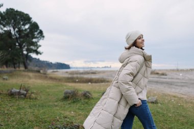 Açık hava sahnesi, yastıklı montlu şık bir kadın kıyıda geziniyor, rüzgarlı kıyı şeridi, kış tarzı ve sakin deniz ufku görünüyor.