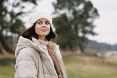Açık havada pofuduk ceketli bir kadın portresi ve örgü bir bere, sakin bir ruh hali, uzak tarlalar arasında doğal manzara, hafif bir esinti ve rahat moda havası.