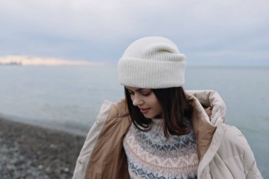 Sahilde bereli ve rahat bir palto giyen bir kadının portresi, kış havası, bulutlu gökyüzü, sakin deniz ve huzurlu kıyı şeridi..