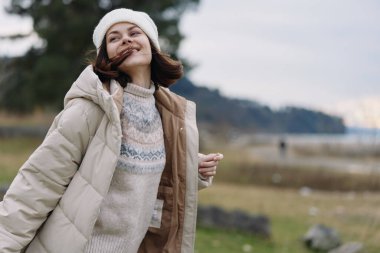 Açık hava sahnesinde sıcacık ceketli ve bereli gülümseyen bir kadın suyun, tepelerin ve yumuşak ışığın altında çıplak ağaçların yanında serin bir manzarada poz veriyor.