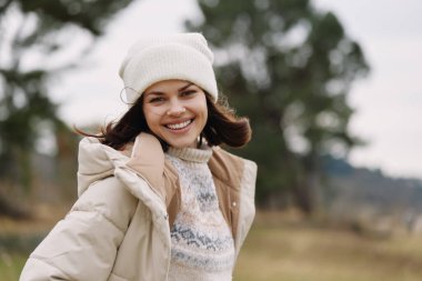 Açık bir alanda ağaçlarla birlikte duran, rahat, ferah ve serin havada mevsimsel tarzını ifade eden, rahat örgü ören, ceketli ve bereli bir kadının açık hava portresi..