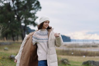 Açık hava kış sahnesinde palto giyen, kazak ören, bereli, rahat poz veren, zahmetsiz bir moda ve sakin bir sonbahar havası yakalayan bir kadın.