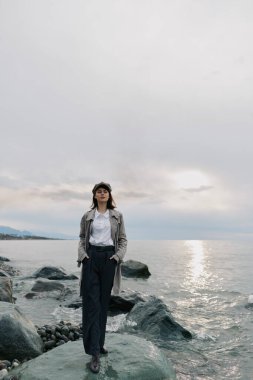 Kendinden emin genç bir kadın bulutlu gökyüzünün altında kayalık sahillerde poz veriyor. Barışçıl doğal ortam bir sakinlik ve yalnızlık havası yaratır..