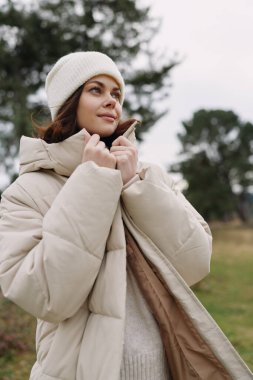 Açık havada bej ceketli ve bereli bir kadının portresi, yumuşak kış ışığı, rahat bir poz, modaya uygun bir dış giysi doğada sıcaklık ve rahatlık sergiliyor.