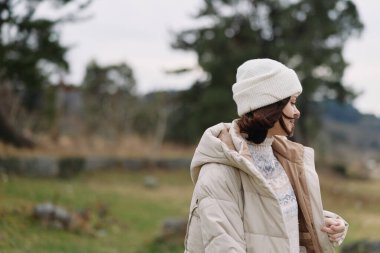 Sıcak bir palto ve beyaz bir bere giyen, sakin bir kırsal alanda göl, ağaçlar ve açık alanlar olan bir kadının sıradan dış portresi.