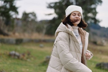 Sıcak giyimli genç bir kadın kırsal alanda temiz havadan, rüzgardan saçlarda ve yumuşak gülümsemelerden, sessiz tonlardan ve açık alanlardan hoşlanır..