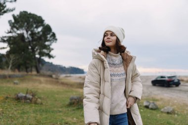 Açık hava sahnesi, kalın bir palto ve örgü süveter giyen şık bir kadın, bereli yumuşak ışık altında manzaralı bir alanda yürüyor, sıcaklık, tarz ve rahat moda çağrıştırıyor.