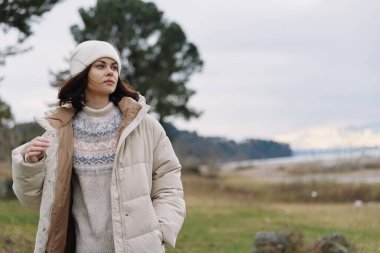 Hava bulutlu bir gökyüzünün, sakin bir ortamın ve şık bir kırsal bölgenin altında kaban giyen ve geniş bir alanda süveter ören bir kadınla sıradan bir açık hava sahnesi.