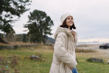 Sıcak bir palto ve şapka giyen gülümseyen bir kadınla sıradan bir açık hava sahnesi, ağaçlı doğal manzara, hayat tarzı fotoğrafçılığı için meltem ve sakin bir gün ışığı.