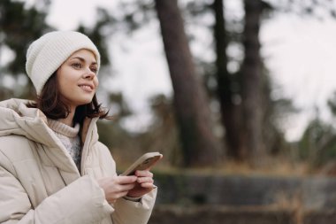 Telefonlu bir kadının açık hava portresi, kalın bir ceket ve bere giymiş, gündelik bir stil sergiliyor, sessiz bir park sahnesi, hafif bir ışık, ve hareketli bir cihazla uğraşırken özenli bir ifade.