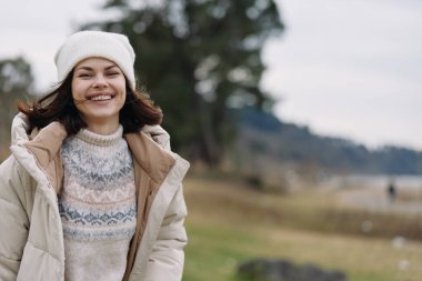 Kışlık ceketli ve bereli gülümseyen bir kadının açık hava portresi, yumuşak ışık, günlük kıyafet ve kentsel arka plan bulanıklığı ve hafif rüzgar ve neşeli ruh haliyle doğal bir manzarada rahat bir poz.
