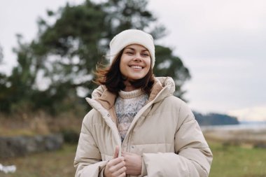 Kışlık ceketli ve bereli gülümseyen bir kadının portresi. Arka planda ağaçlar ve su olan bir tarlada duruyor. Atmosferik ve sakin bir manzara..