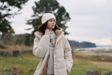 Kabarık ceketli ve rahat kazaklı yalnız bir kadın çimenli tarlaların, bulutlu gökyüzünün, sakin atmosferin, ince moda detaylarının ve cömert negatif alanların arasında poz verdi..